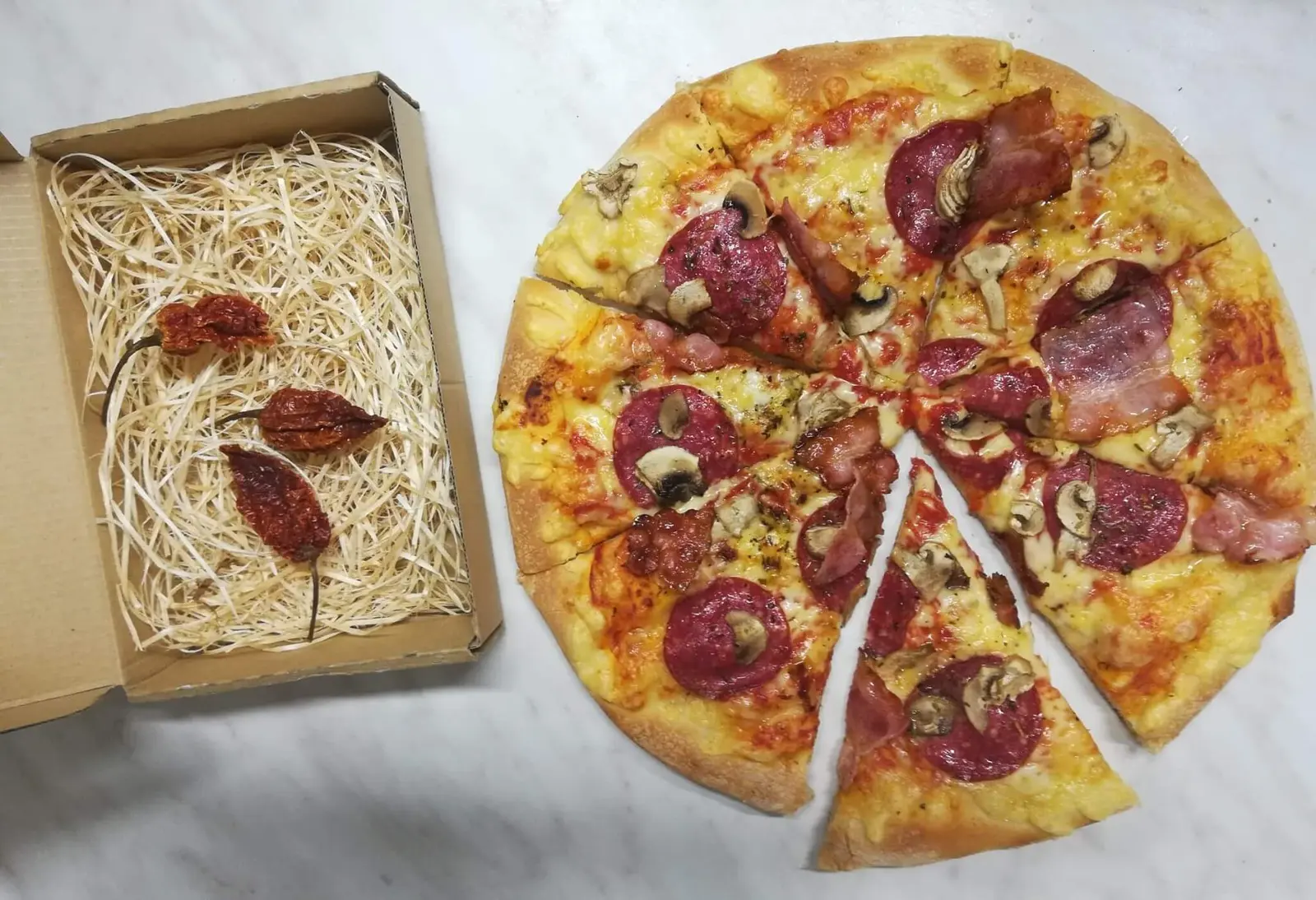 Pokrojona pizza z salami, boczkiem i pieczarkami obok pudełka z suszonymi papryczkami chili