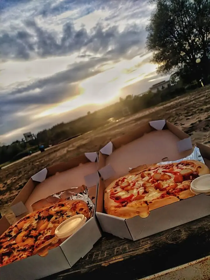 Dwie pizze w otwartych pudełkach na drewnianym stole, w tle piasek, drzewa i zachód słońca