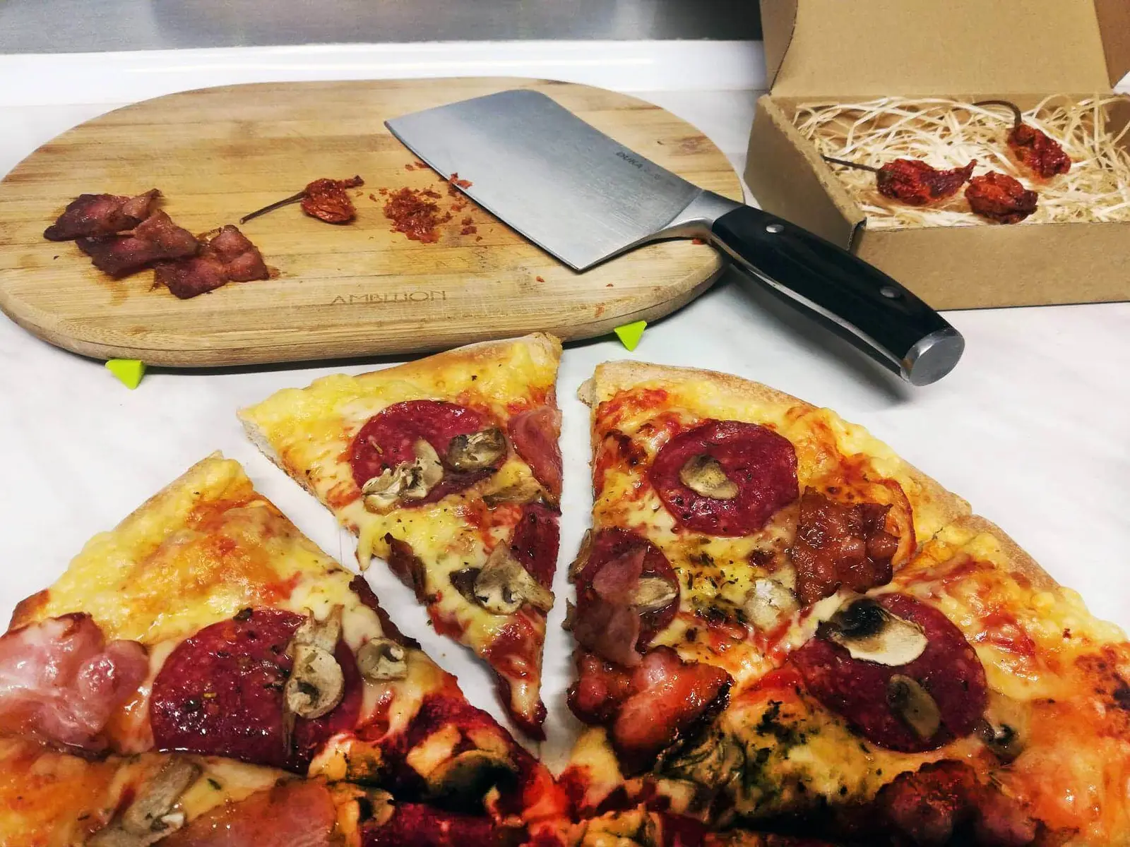Kawałki pizzy z salami i pieczarkami, w tle deska z tasakiem i suszone papryczki chili